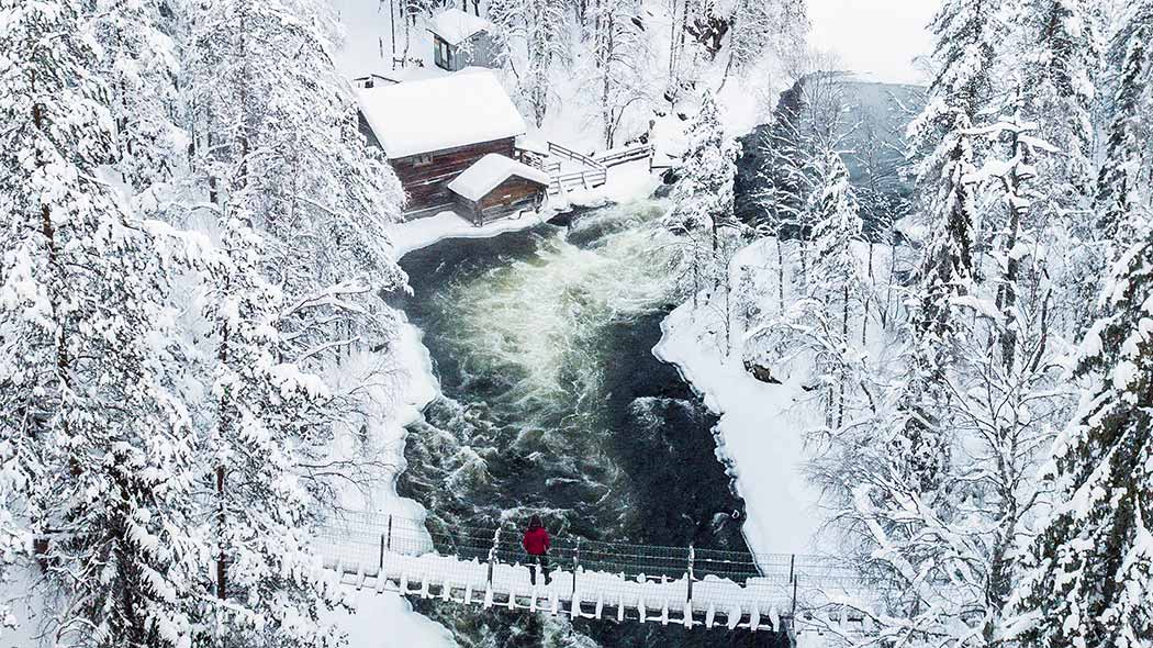 悬索桥上有个人正在观望着白雪皑皑的河流和一座庞大的木质结构建造的磨坊景观。